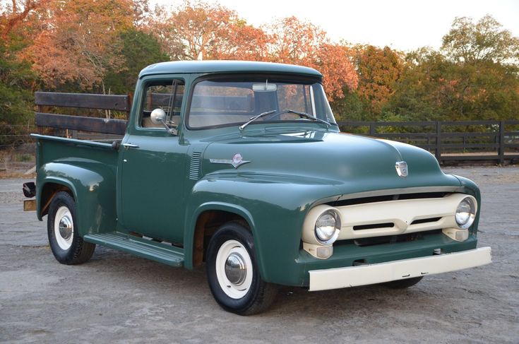 an old green truck parked in a parking lot next to a wooden fence and trees