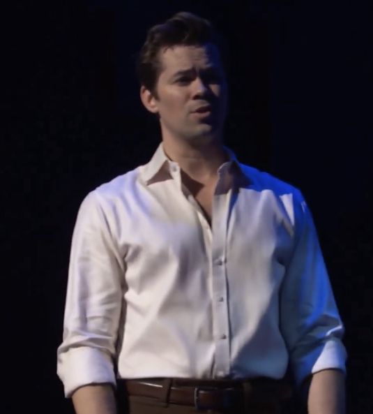 a man in a white shirt and brown pants standing on stage with his hands in his pockets