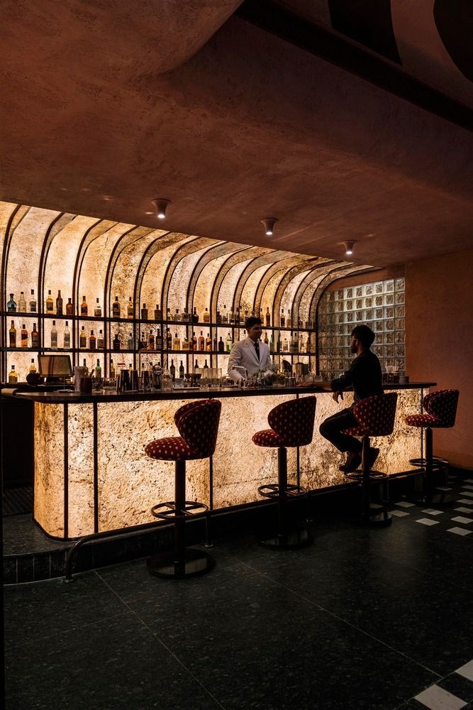 a man sitting at a bar with lots of bottles on the wall behind him and stools in front of it