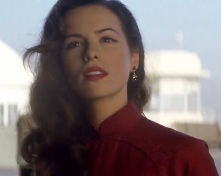 a woman with long dark hair wearing a red shirt and earrings, standing in front of a white building