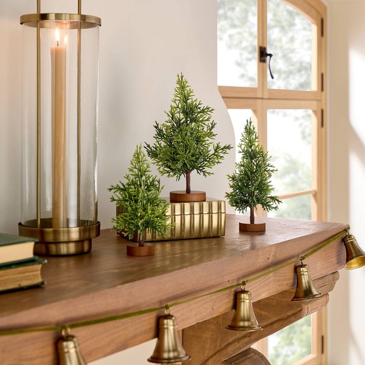 three small trees are on top of a mantle in front of a window with two brass bells