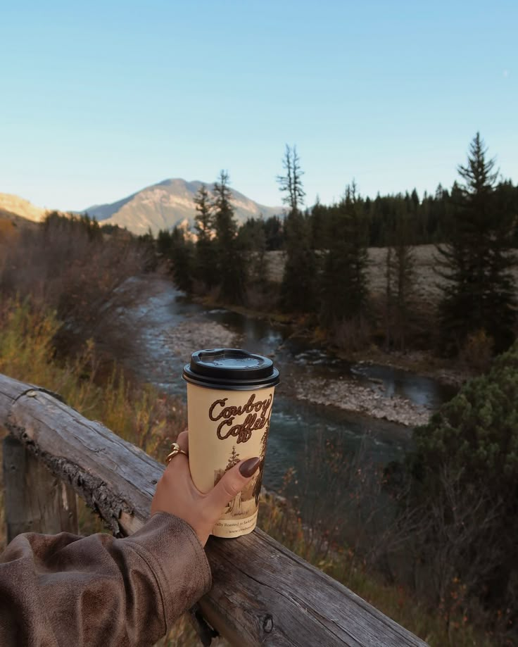 a person holding up a coffee cup on top of a wooden railing near a river