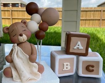 a teddy bear is sitting on a cake with balloons in the shape of letters and blocks