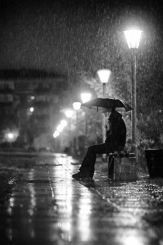 a person sitting on a bench under an umbrella in the rain at night with street lights