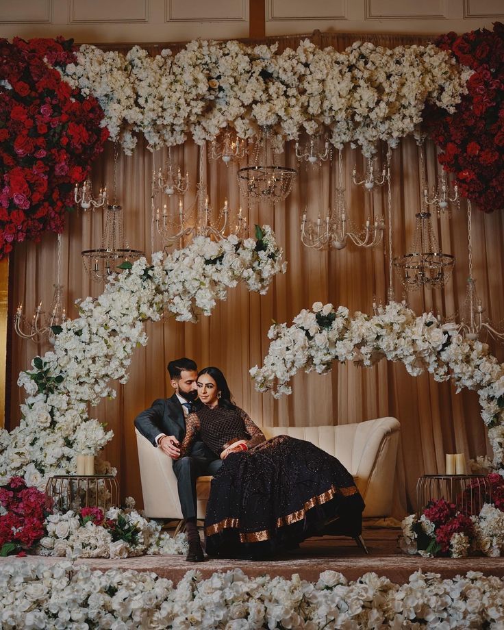 a man and woman sitting on a couch in front of a floral wall with chandeliers