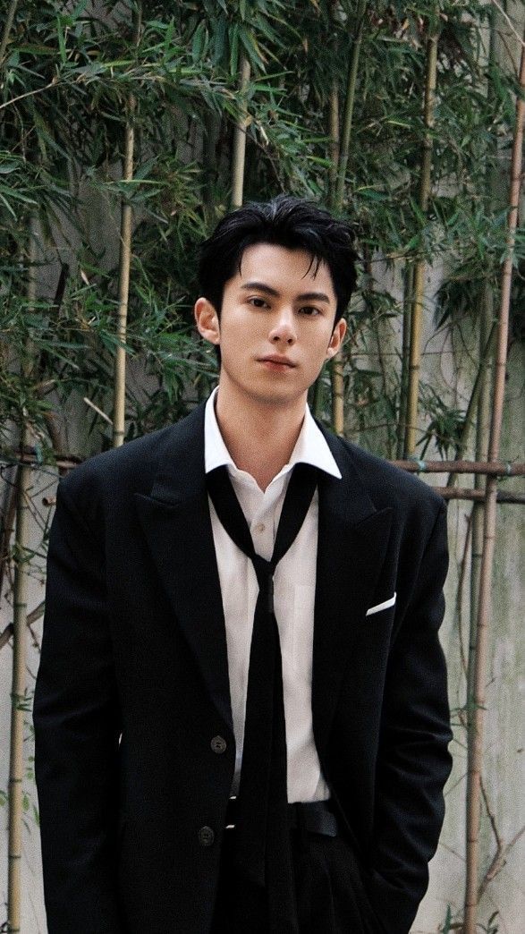 a young man wearing a suit and tie standing in front of a bamboo tree with his hands in his pockets