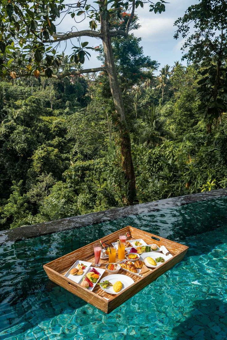 a wooden tray filled with food sitting on top of a swimming pool next to trees