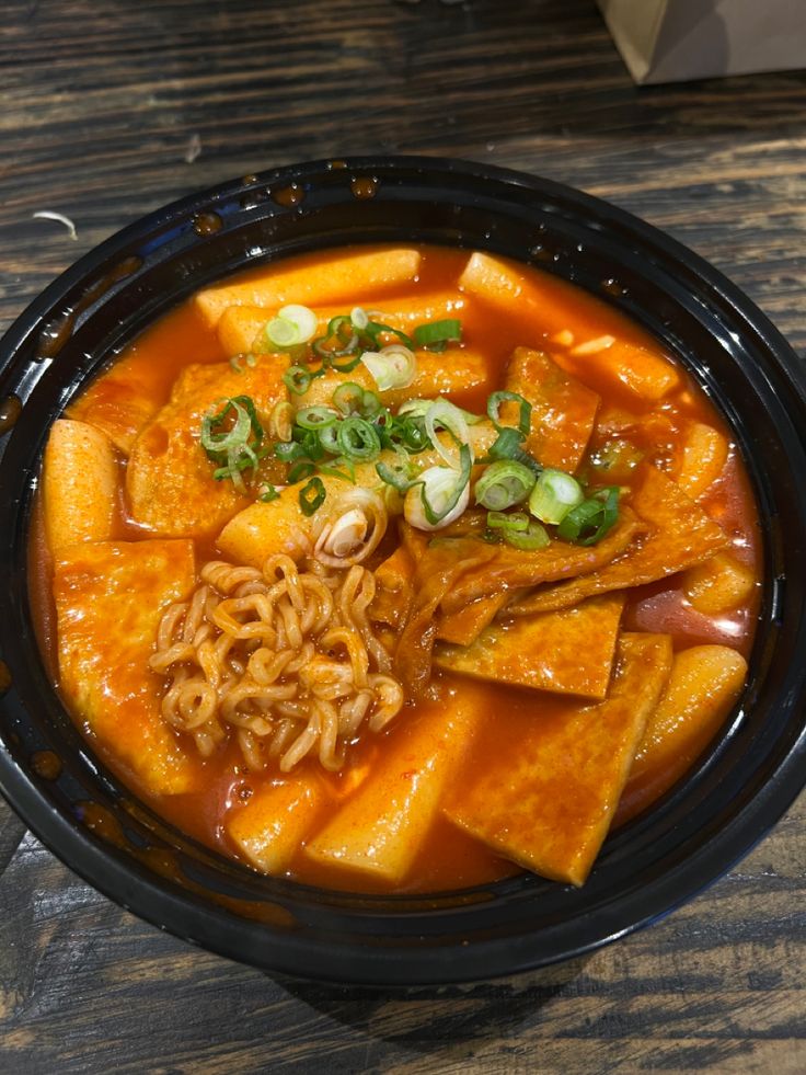 a black bowl filled with noodles and vegetables on top of a wooden table next to a cup of coffee