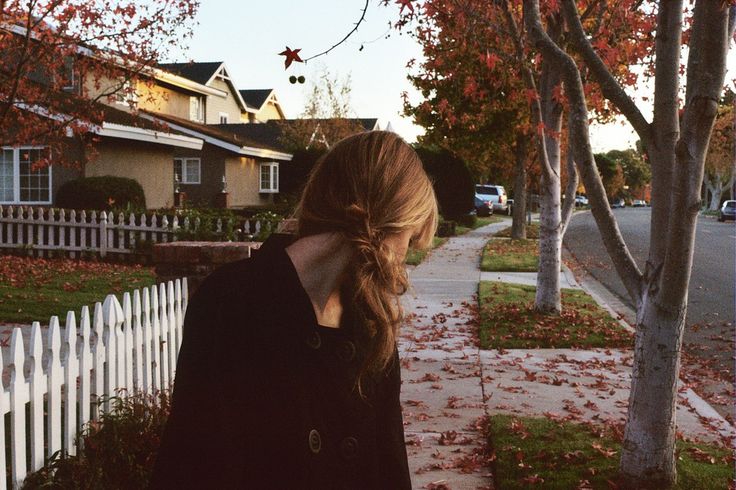 a woman standing on the sidewalk in front of a white picket fence and trees with red leaves