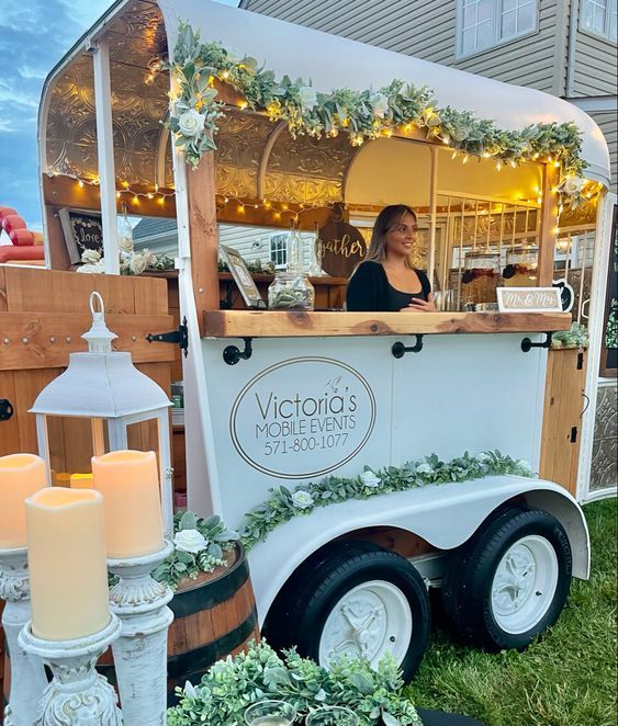 a woman standing in front of a food truck with candles on the side and greenery around it