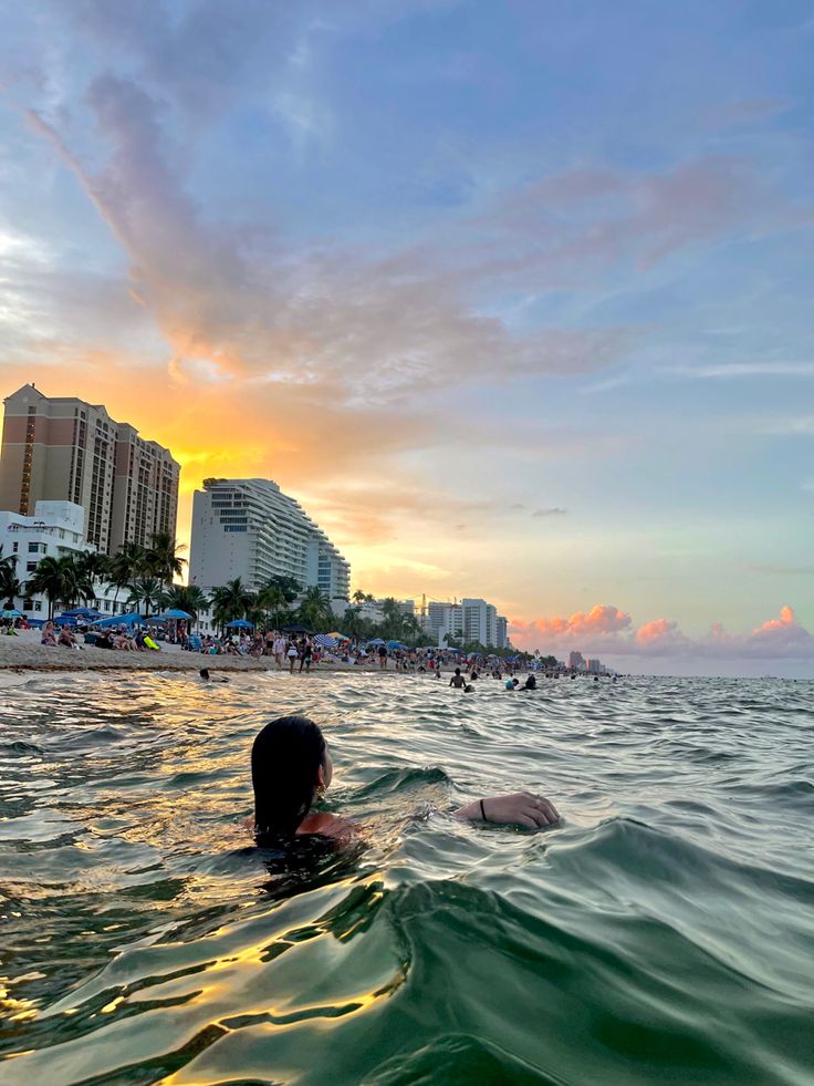 a woman swimming in the ocean at sunset with buildings in the background and people on the beach