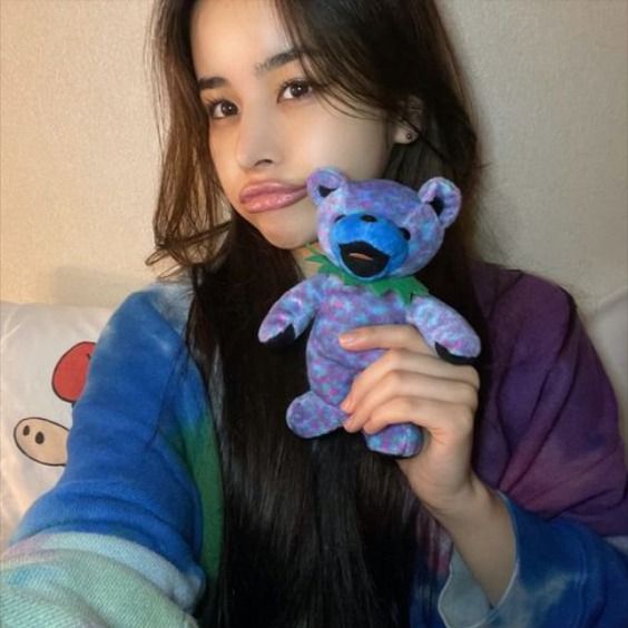 a woman holding a blue teddy bear in her right hand and looking at the camera