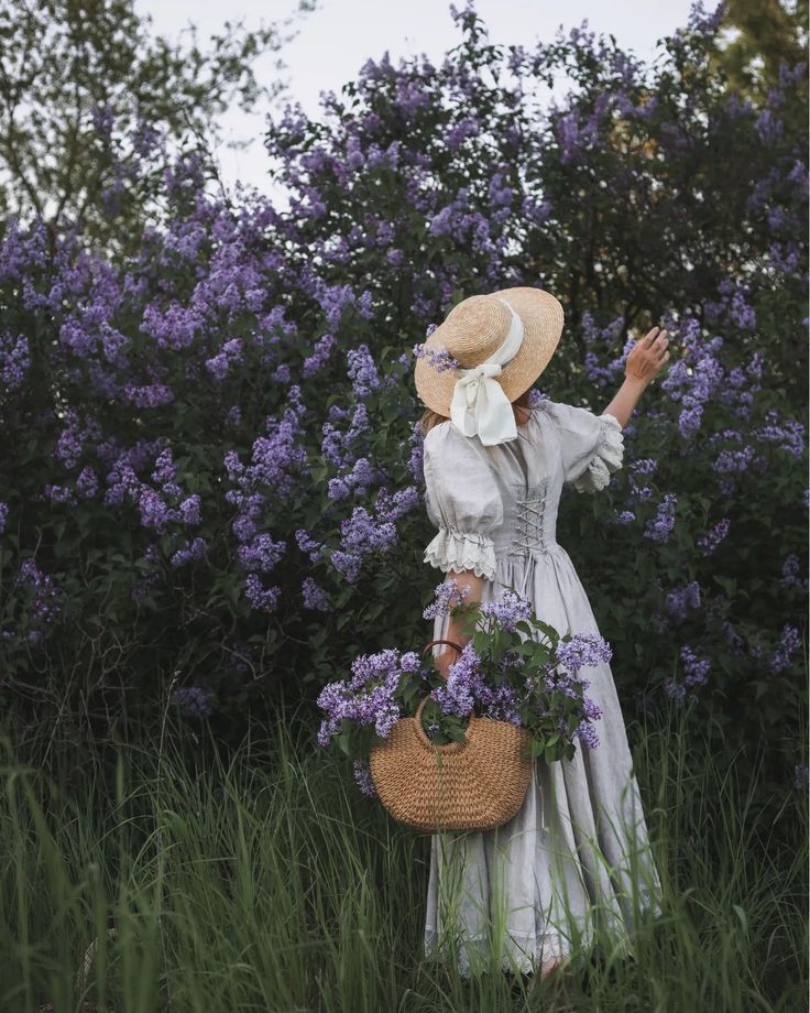 a woman in a white dress and straw hat holding a basket full of purple flowers