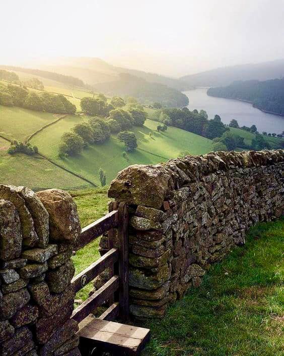 a stone wall and wooden bench on the side of a grassy hill overlooking a body of water