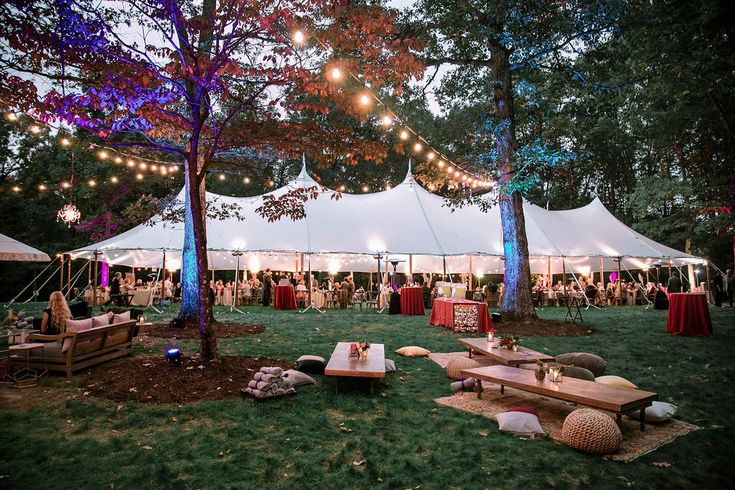 a group of people sitting under tents in the grass with lights strung from trees above them