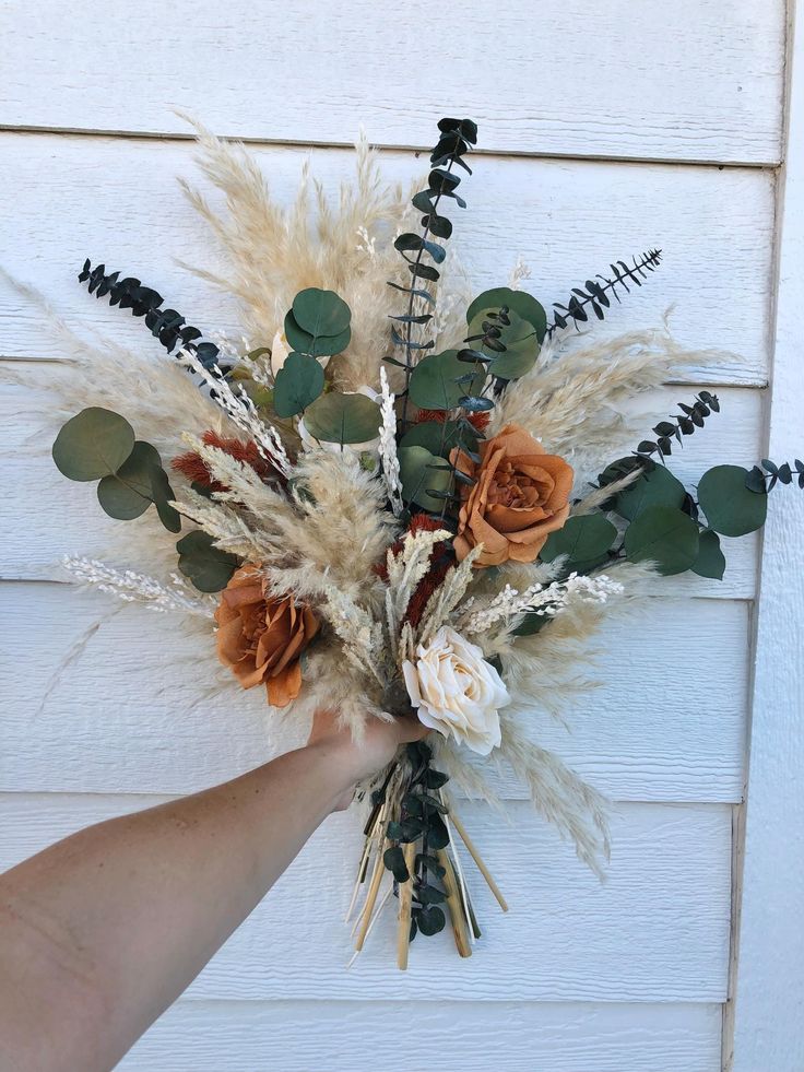 a hand holding a bouquet of dried flowers and greenery in front of a white wall