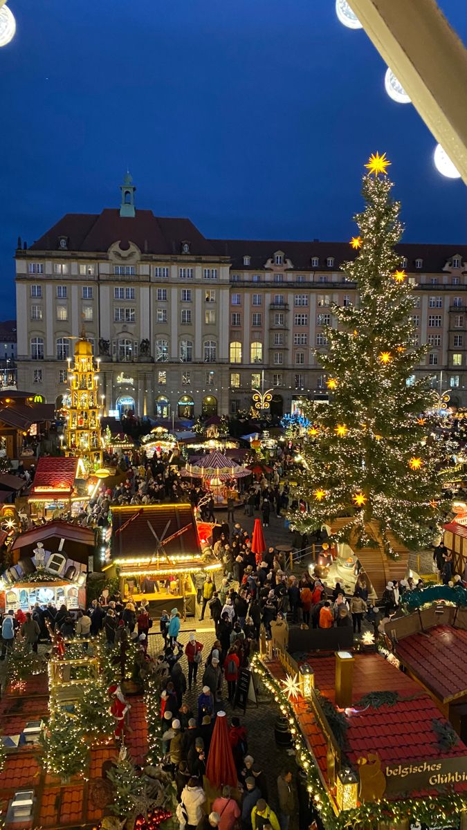 an outdoor christmas market is lit up at night with lights on the trees and people walking around