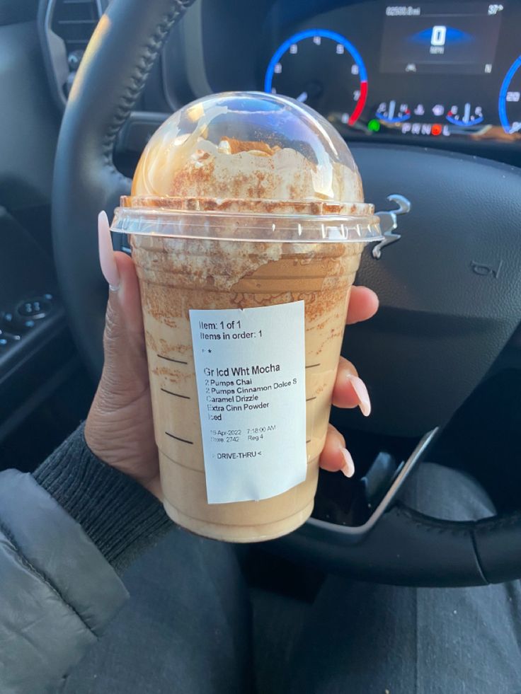 a person holding up a drink in front of a car steering wheel and dashboard gauges