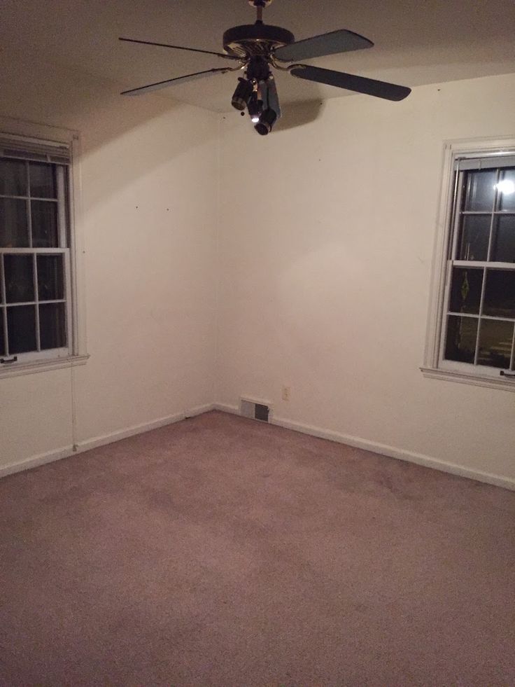 an empty room with two windows and a ceiling fan in the corner on the far wall