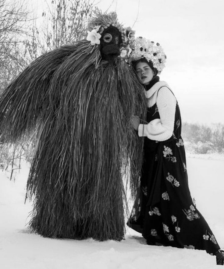 a woman is standing in the snow next to a tall grass monster with flowers on it's head