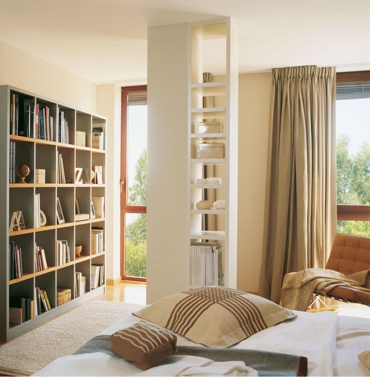 a bedroom with a book shelf, chair and bed in front of a large window