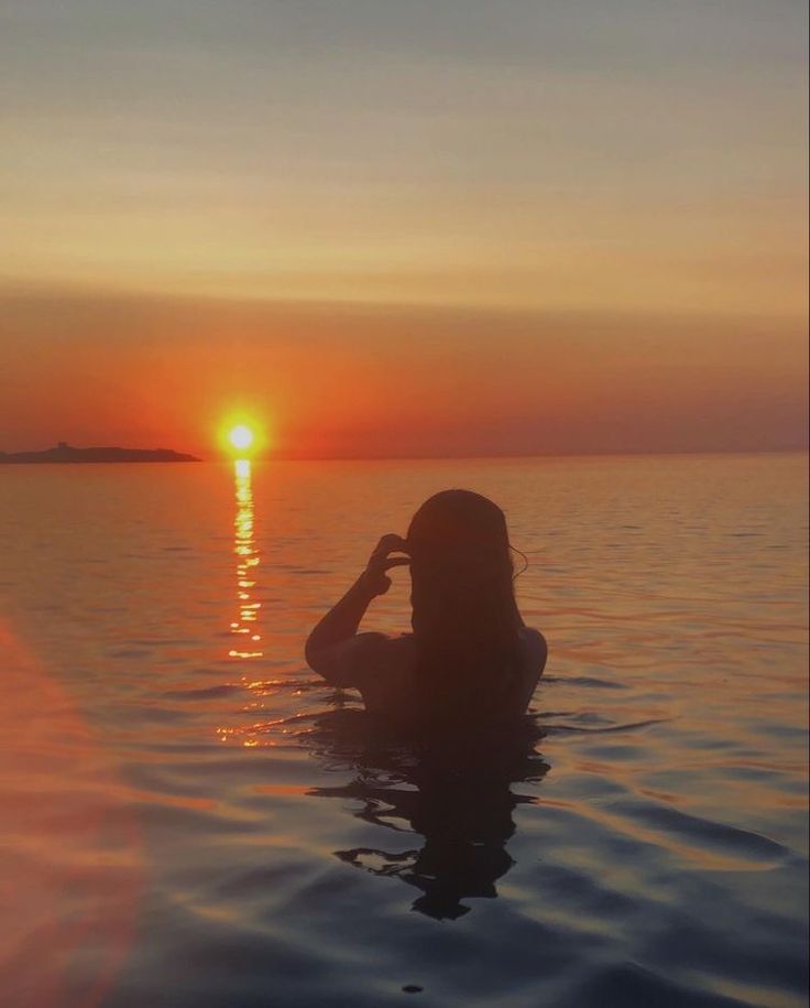 a woman sitting in the water at sunset with her hands on her head, looking out to sea