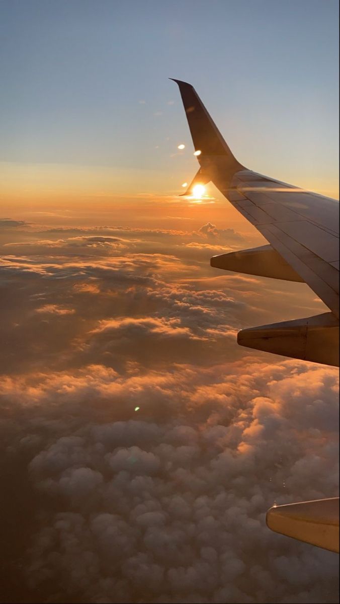 the wing of an airplane as it flies above the clouds at sunset or sunrise, with the sun peeking through the clouds