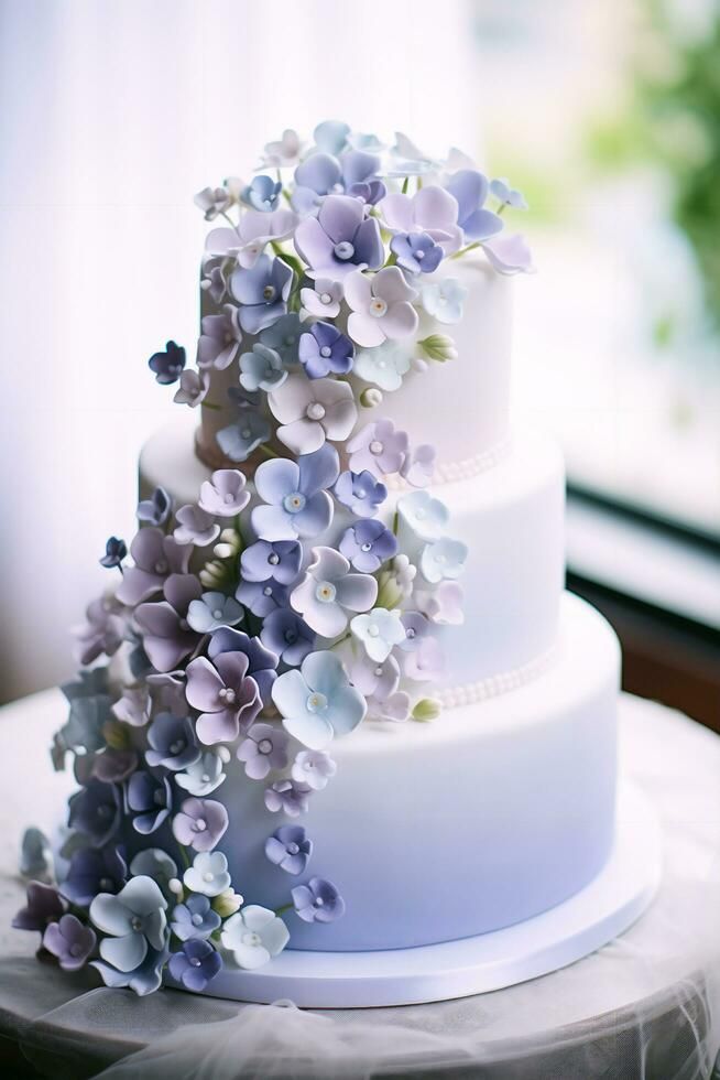 a three tiered white cake with purple flowers on the top and bottom, sitting on a table in front of a window