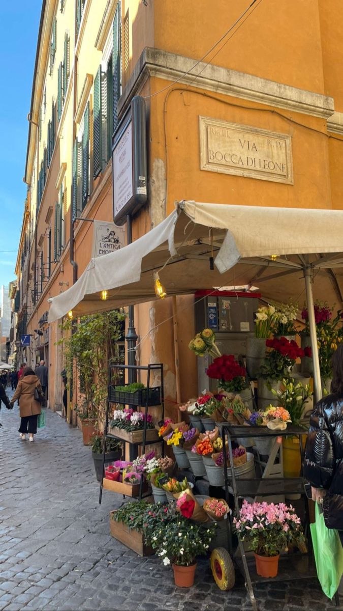 an outdoor flower shop on the side of a street with people walking by it and some buildings in the background