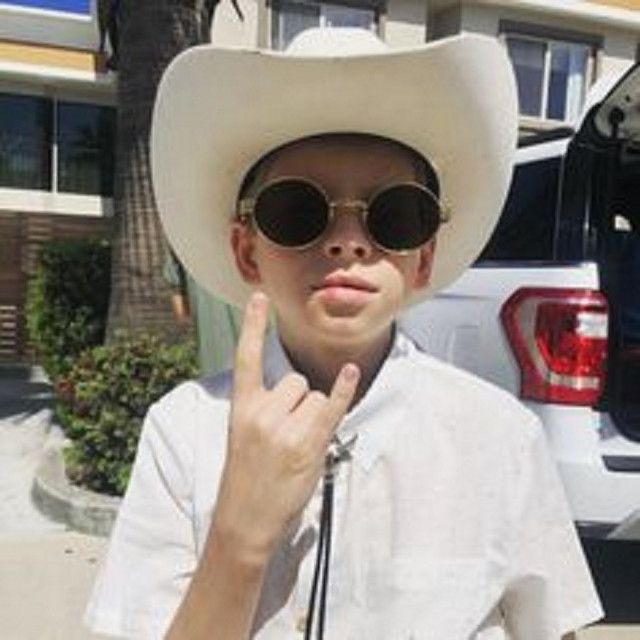 a young boy wearing a white cowboy hat and sunglasses making the peace sign with his fingers