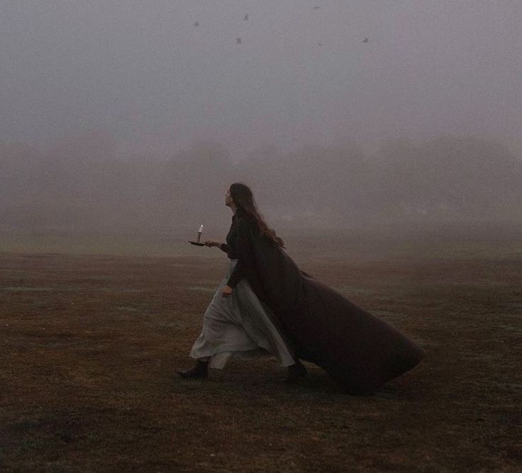 a woman walking in the fog with a long coat over her shoulders and a candle in her hand