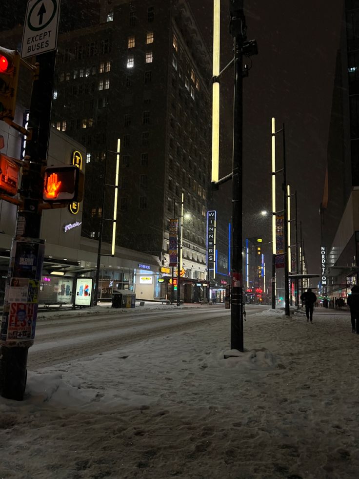 people are walking in the snow on a city street at night with tall buildings and traffic lights