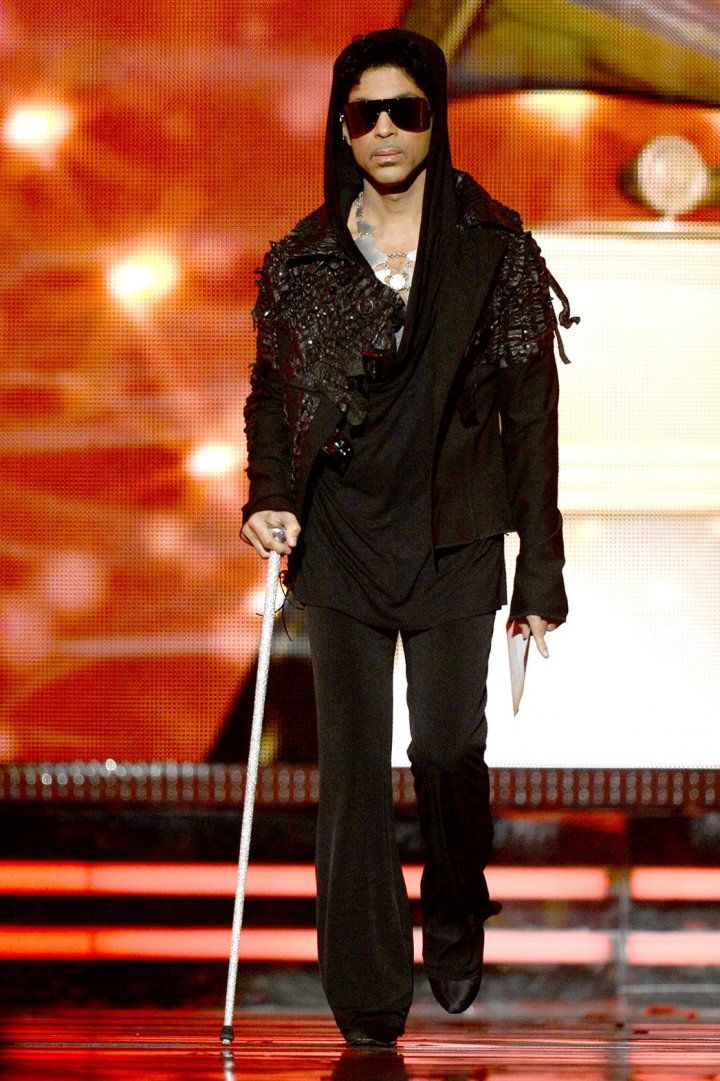 the man is walking down the runway with a cane in his hand and wearing sunglasses