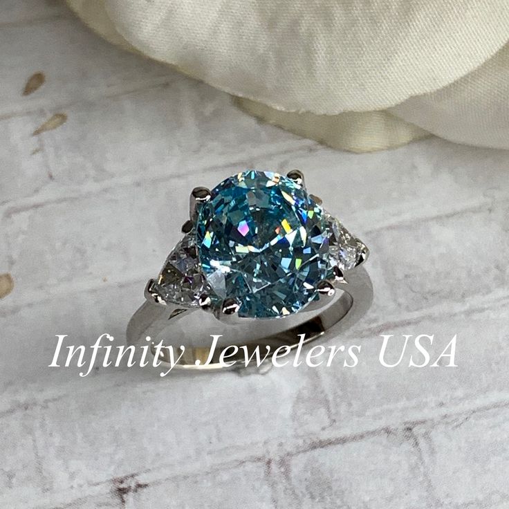 "The ring pictured is a simulated aquamarine with simulated diamonds #7034 -Approximate total carat weight: 5.00ctw diamond equivalent -Center Stone Size: 11x9mm - approx. 4.20ct diamond equivalent -Center Stone Shape: oval cut -Side Stones Size: 2 Trillions 5x5mm - approximately 0.80ctw diamond equivalent -Gem Type: simulated diamond -Stone Clarity: VVS1 -Stone Color: light blue center stone -Moh's Scale: 8.5 hardness -Metal Type and Purity: 14k white gold -Setting: 4 prong tulip head -Stock Ri Oval Aquamarine Diamond Ring With Prong Setting, Light Blue Oval Ring With Prong Setting, Light Blue Diamond Ring For Anniversaries, Anniversary Light Blue Aquamarine Diamond Ring, Oval Light Blue Promise Ring, Light Blue Oval Promise Ring, Anniversary Light Blue Diamond Ring, Anniversary Aquamarine Diamond Ring With Halo Setting, Oval Aquamarine Ring With Diamond Accents