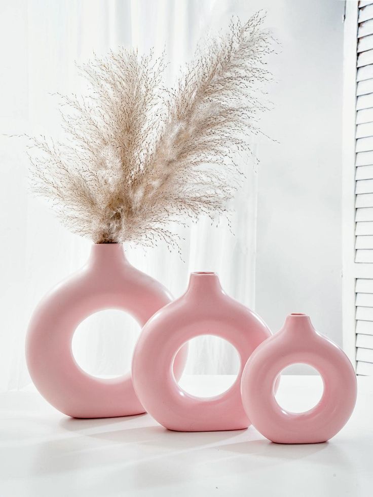 three pink vases sitting next to each other on a white surface with a plant in the middle