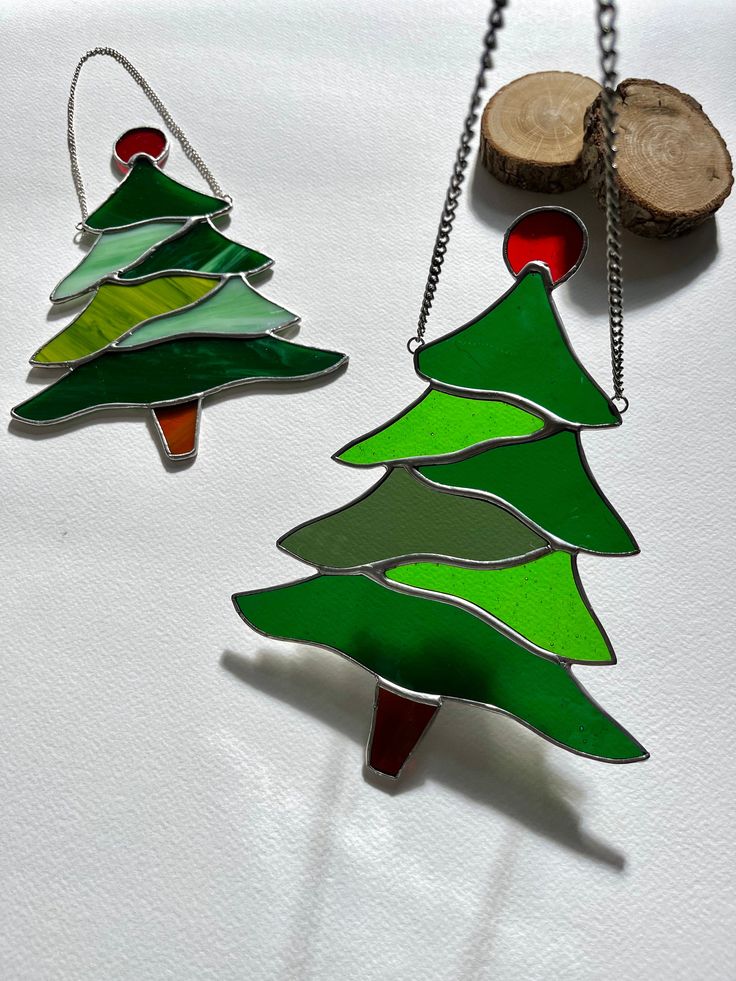 two stained glass christmas trees hanging from chains next to a piece of wood on a white surface