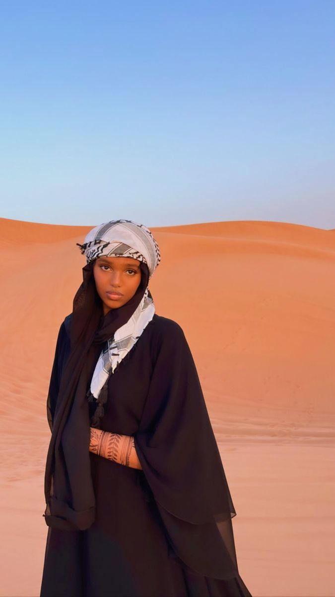 a woman standing in the desert wearing a black dress and headdress with her hands on her hips