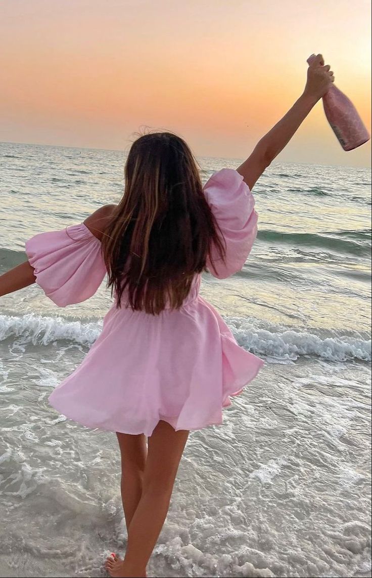 a girl in a pink dress is walking on the beach with her arms raised up