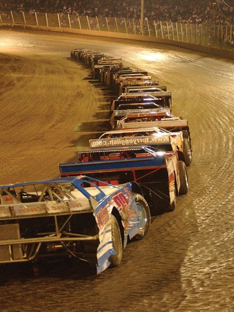 a row of boats sitting on top of a dirt covered field at night, with people watching from the stands