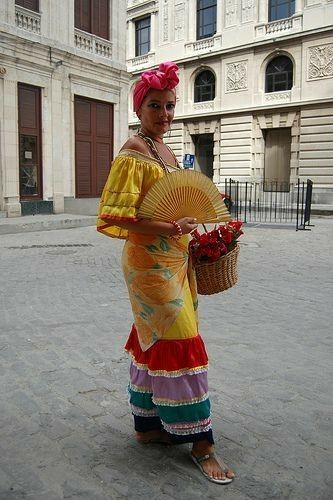 a woman in colorful dress holding a fan and flowers on the street with buildings behind her