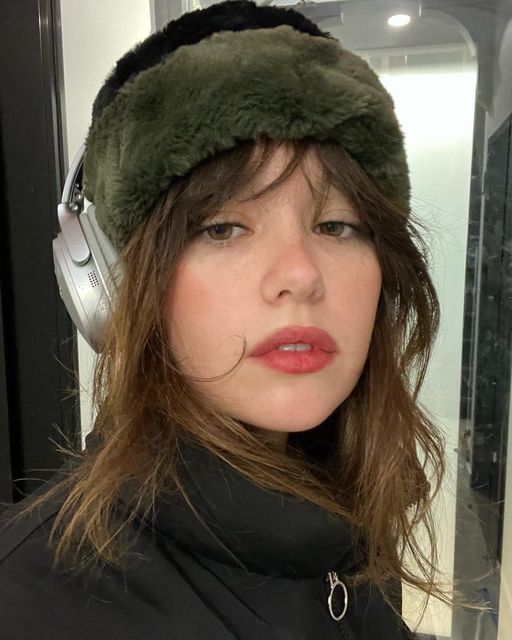 a woman with headphones on and wearing a green fur hat in front of a mirror