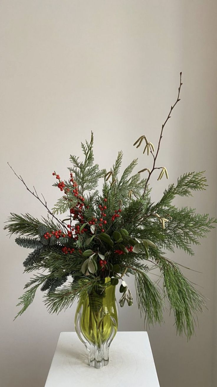 a green vase filled with red berries and greenery on top of a white table