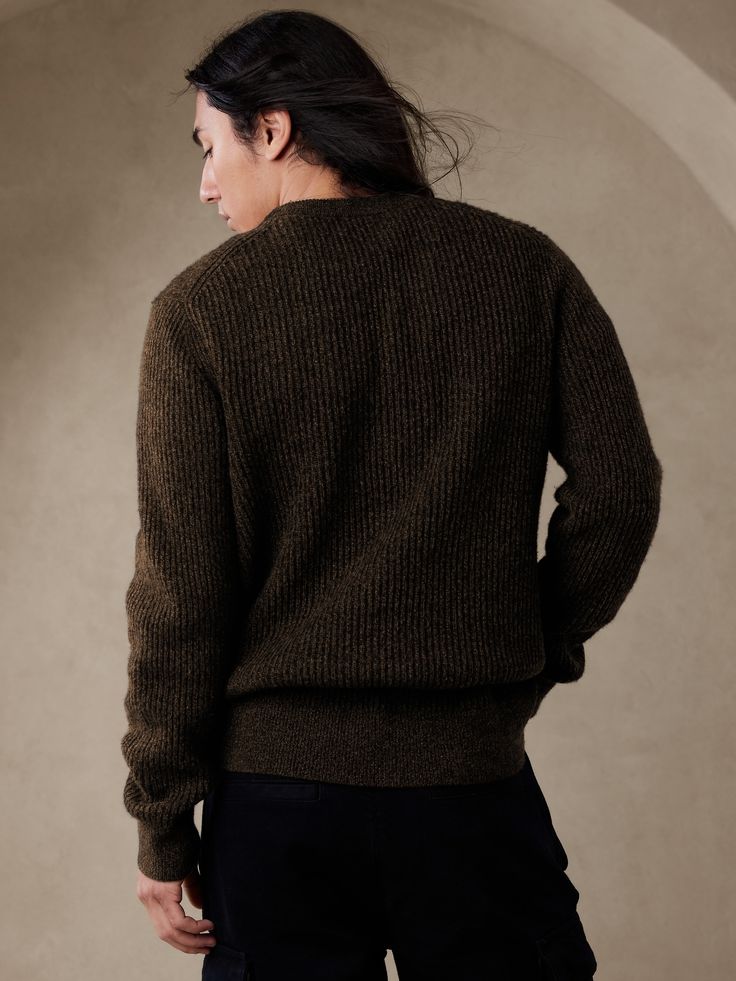 a woman standing in front of a wall with her back turned to the camera, wearing a brown sweater and black pants