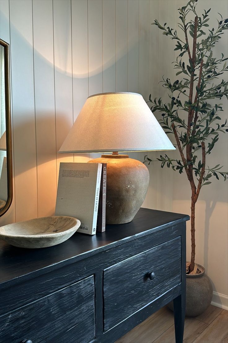 a lamp on top of a dresser next to a mirror and vase with a tree in it