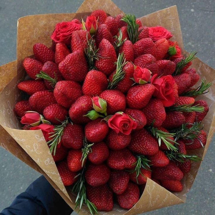 a bouquet of strawberries with red roses and greenery on top is held in someone's hand