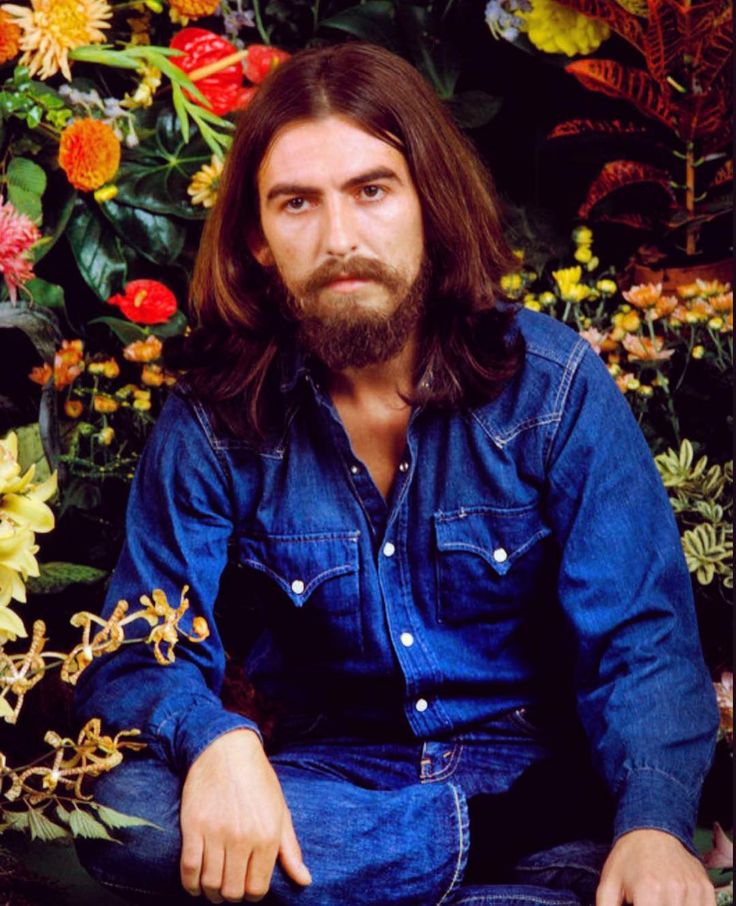 a man with long hair and beard sitting on the ground in front of colorful flowers