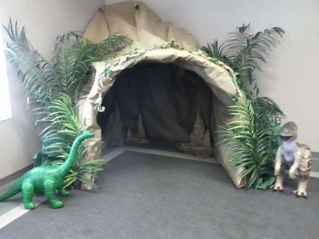 two toy dinosaurs are sitting in front of a fake cave with plants and rocks on the floor