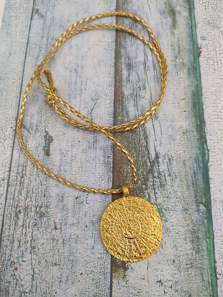 Stunning large Gold Phaistos disk pendant with plaited gold cord (and length of black 2mm cotton cord) This pendant can become a fascinating focal point of conversation and will look stunning in Summer or Winter.  The original Phaistos Disc is on display at the archaeological museum of Heraklion. It is a disk of fired clay from the Minoan palace of Phaistos on the island of Crete, dating to the middle or late Minoan Bronze Age (which is the second millennium BC). The disk is about 15 cm diameter Spiritual Brass Coin Necklace With Round Pendant, Handmade Brass Spiritual Coin Necklace, Hammered Medallion Amulet Jewelry, Round Large Pendant Coin Necklace Amulet Style, Amulet Style Coin Necklace With Large Round Pendant, Round Amulet Style Coin Necklace With Large Pendant, Round Coin Necklace With Large Amulet Pendant, Round Coin Necklace With Large Pendant Styled As Amulet, Handmade Yellow Gold Medallion Necklace In Brass