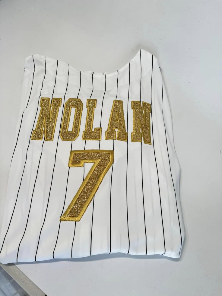 Stand out at the game with our Custom Pinstripe Baseball Jersey. This jersey features intricate embroidery and glitter applique, making it truly one-of-a-kind. Show off your unique style and personality with this playful and fun addition to your baseball gear. (And impress your teams, too!) Pricing includes one name on the back and one number, additional customization can be added, please ask for details on pricing! PLEASE NOTE the name and number, as well as glitter color in the notes! Product Details Feature: Pad print label Set-in sleeves Full-button jersey Double-needle hemmed sleeves and fishtail bottom. Sizing - See attached photo Fabric : 100% polyester smooth pinstripe knit / Wicks moisture away from the body Fit - Classic unisex fit Made to order: one week ship time White Tops With Three Stripes And Baseball Collar, Collegiate Striped Tops For Game Day, Striped Collegiate Tops For Game Day, Sporty Baseball Jersey With Contrast Stripes, Game Day Striped Baseball Jersey With Letter Print, Striped Baseball Jersey With Letter Print For Game Day, Varsity Baseball Jersey With Three Stripes For Sports Season, Varsity Baseball Jersey With Contrast Stripes, Team-colored Tops With Three Stripes For Baseball Season