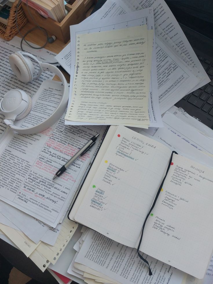 many books and headphones are on top of the desk covered with papers that have been stacked together